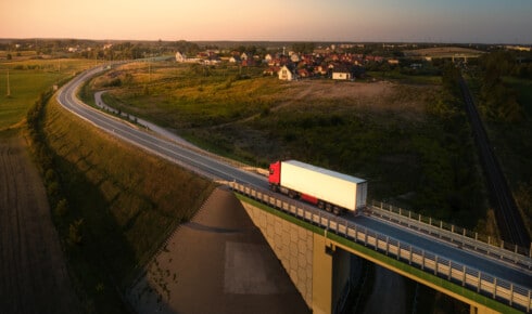 Luftaufnahme eines Lastwagens auf einer Landstra&szlig;e bei Sonnenuntergang, mit einem Dorf und einer Br&uuml;cke in einer nordischen Landschaft &ndash; sinnbildlich f&uuml;r einen Fuhrpark, der mit erneuerbarem Diesel f&auml;hrt