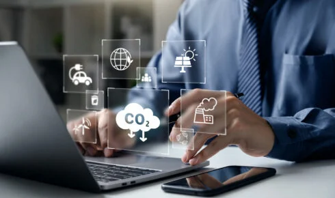 Eine Fachperson arbeitet an einem Laptop, umgeben von digitalen Symbolen für CO2-Emissionen, eine Fabrik, erneuerbare Energie und ein Elektrofahrzeug – sinnbildlich für die digitale Nachverfolgung hinter dem betrieblichen Emissions-Reporting