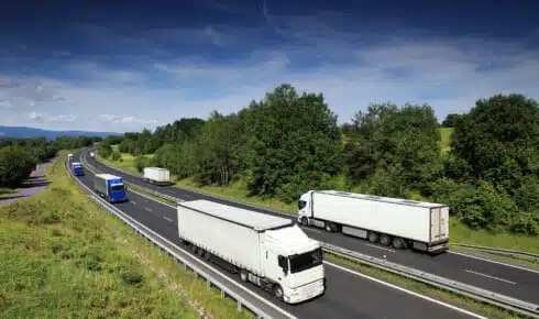 Mehrere schwere Lkw fahren auf einer mehrspurigen Autobahn durch gr&uuml;nes H&uuml;gelland &ndash; sinnbildlich f&uuml;r den Stra&szlig;eng&uuml;terverkehr, der den EU-Klimavorschriften im Transport unterliegt